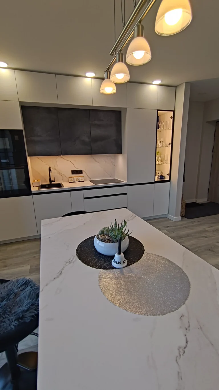 Vakarinis Virtuvės Apšvietimas Kaune Kreves Prospekte Pilna Vidaus Apdaila Balta Virtuvė Su Tamsia Viršutine Sekcija Ir šiltai Apšviestas Baro Sala